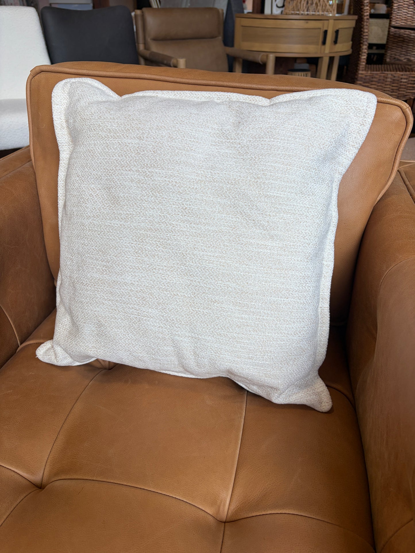 A square off-white textured throw pillow with a white piped edge, resting on a brown leather sofa.