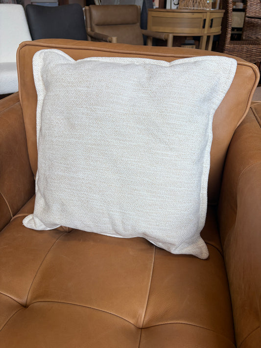 A square off-white textured throw pillow with a white piped edge, resting on a brown leather sofa.