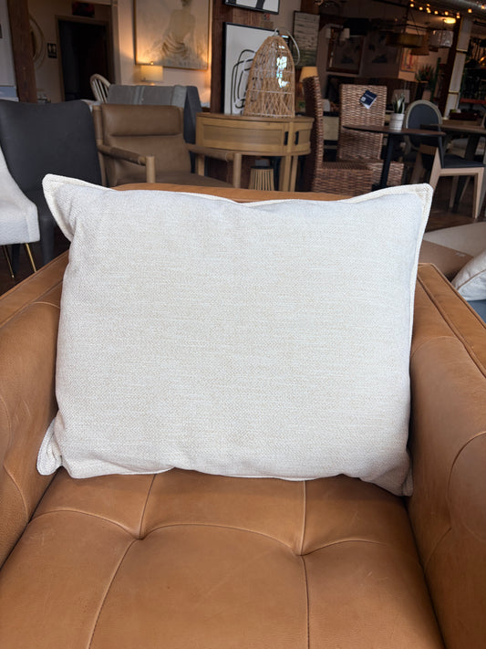 A square off-white textured throw pillow resting on a tan leather tufted armchair in a furniture showroom.